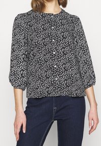 Blouse noire à motif floral blanc, col rond et fermeture à boutons. Manches longues bouffantes avec poignets froncés. Coupe décontractée.