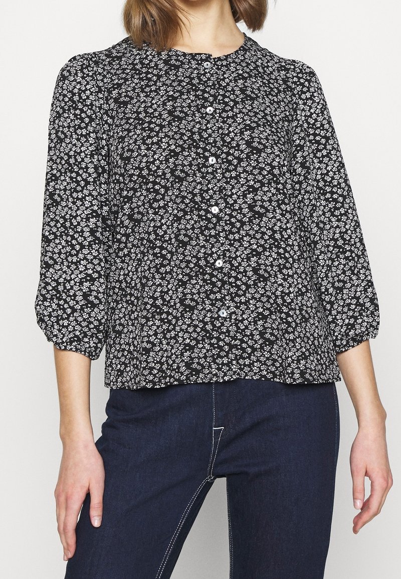 Blouse noire à motif floral blanc, col rond et fermeture à boutons. Manches longues bouffantes avec poignets froncés. Coupe décontractée.