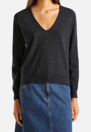 Kobieta ubrana w ciemnoszary sweter z długim rękawem i dekoltem w serek oraz niebieską spódnicę jeansową, stojąca z rękami swobodnie wzdłuż ciała.