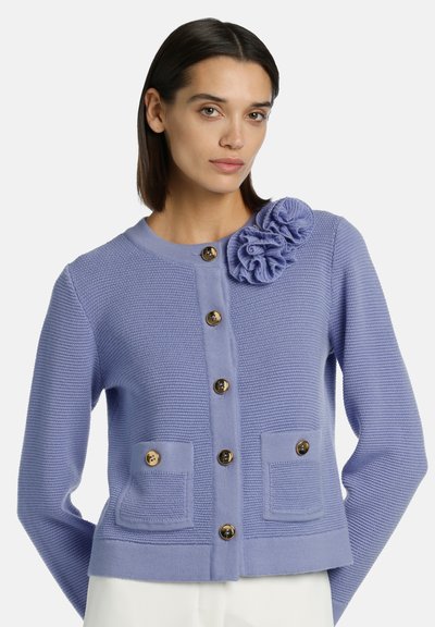 Cardigan de color lila claro con un tejido texturizado, que presenta un adorno de flor con volantes, botones dorados y dos bolsillos frontales.