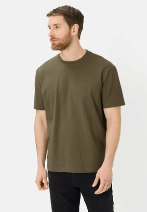 Mężczyzna z brodą, ubrany w gładką oliwkowozieloną koszulkę z krótkim rękawem i czarne spodnie, patrzący w lewo na białym tle.