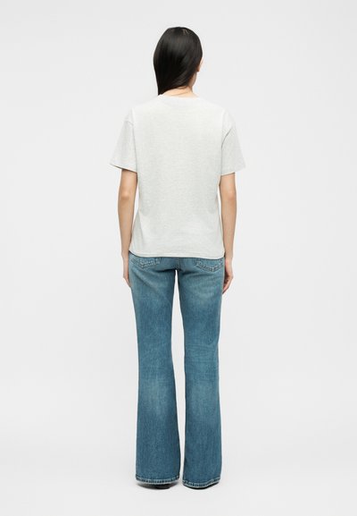 Γυναίκα με μακριά μαύρα μαλλιά, γυρισμένη από πίσω, φοράει απλό ανοιχτό γκρι κοντομάνικο μπλουζάκι και μπλε τζιν bootcut, μπροστά σε λευκό φόντο.