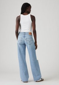 Jeans en denim bleu clair à jambes larges avec une taille haute, des poches latérales et une patch en cuir beige au dos. Portés avec un débardeur blanc et des sandales.
