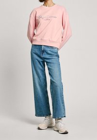 Sudadera de algodón rosa claro con texto de logo blanco, combinada con jeans de mezclilla azul de pierna ancha y zapatillas deportivas beige.