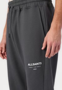 Graue Jogginghose mit elastischem Bund, Seitentaschen und weißem Logo-Text auf dem linken Oberschenkel. Weicher, glatter Stoff.