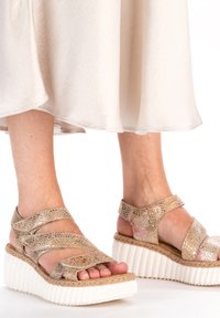 Sandales compensées avec une semelle en jute tressé, partie supérieure dorée métallisée en matériau semblable au cuir, avec un design à brides croisées et bout ouvert.