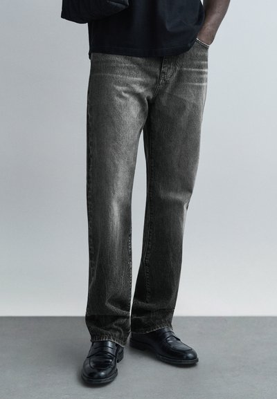Μαύρο τζιν από denim με ξεθωριασμένη υφή, κανονική ευθεία γραμμή και σχεδιασμό με πέντε τσέπες, σε συνδυασμό με μαύρα παπούτσια. Ορατές είναι οι λεπτές ραφές.