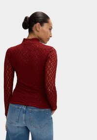 Top in pizzo rosso con maniche lunghe, colletto alto, motivo testurizzato, abbinato a jeans in denim azzurro chiaro, visto da dietro.