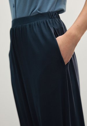 Person wearing dark loose pants with elastic waistband, hand resting inside right pocket, blue shirt tucked in.