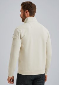 Sudadera de color crema claro con collar alto, que presenta un panel en la parte posterior y un bolsillo funcional en la manga. Tejido suave y texturizado.