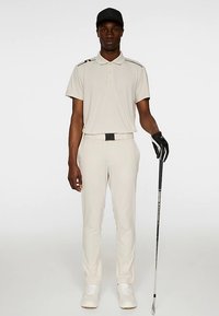 Ljusbeige golfklädsel med en pikétröja med svarta detaljer, skräddarsydda byxor och en svart keps. Golfaren håller i en klubba och bär en handske.