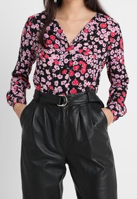 Blusa nera floreale con fiori rosa e rossi, design a scollo a V, maniche lunghe. Indossata con pantaloni in pelle neri a vita alta con fibbia.