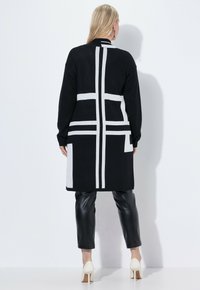 Lång svart cardigan med vitt geometriskt mönster, mjuk textur, öppen fram, i kombination med svarta läderbyxor och vita klackskor.