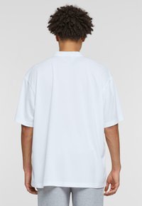 Camiseta blanca de gran tamaño con mangas cortas, cuello redondo y una textura de tela suave, mostrada desde la parte de atrás con pantalones grises.
