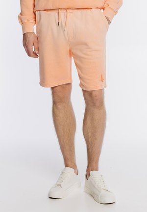 Mężczyzna ubrany w brzoskwiniowe szorty i pasującą bluzę, w białych trampkach, stoi z rękami w kieszeniach na białym tle.