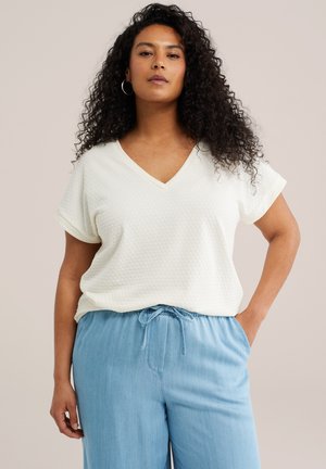 Žena s dlhými kučeravými vlasmi nosí biely krátky rukáv s výstrihom do V a svetlomodré nohavice so šnúrkou, stojí s jednou rukou v vrecku.