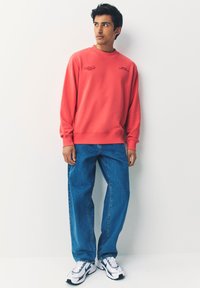 Felpa color corallo con polsini e collo a coste, abbinata a jeans blu a taglio ampio e sneakers bianche con dettagli blu.