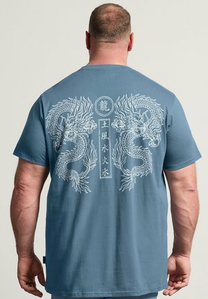 Homme portant un t-shirt bleu avec des motifs de dragons jumeaux blancs et des caractères chinois verticaux dans le dos, debout sur un fond clair.