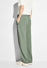 Persoon gekleed in lichtgroene wijde broek, wit T-shirt en beige chunky sneakers, staand met handen in de zakken tegen een witte achtergrond.