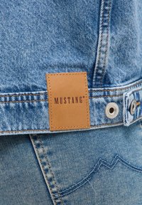 La veste en denim présente une étiquette en cuir marron étiquetée "MUSTANG", avec des accessoires en métal argenté et des coutures contrastées sur un tissu bleu clair.