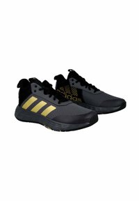 Zapatillas deportivas negras con parte superior de malla gris, tres franjas doradas y acentos negros. Presentan una suela texturizada y un diseño de punta redondeada.