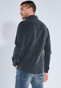 Langärmliges Hemd in Dunkelgrau mit schmalen vertikalen Streifen, aus strukturierter Stoff gefertigt. Verfügt über einen Kragen und Knopfmanschetten. Kombiniert mit blauen Jeans.