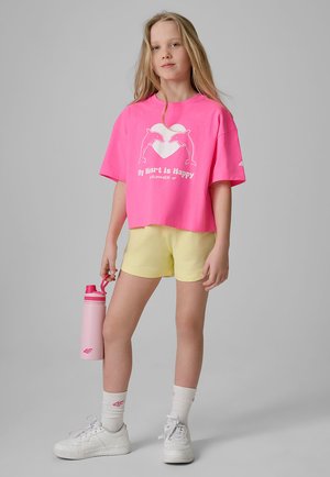 Młoda dziewczyna w różowym T-shircie z delfinami i napisem, żółtych szortach, białych trampkach i skarpetkach trzymająca różową butelkę na wodę na szarym tle.