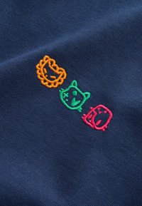 Tre piccole facce di animali da cartone ricamate in arancione, verde e rosso su tessuto blu scuro.