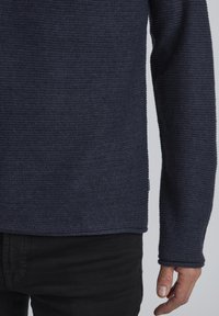 Marineblauer strukturierter Pullover mit geripptem Design, langen Ärmeln und einem leicht lockeren Schnitt. Kombiniert mit schwarzen Hosen, die am unteren Ende sichtbar sind.