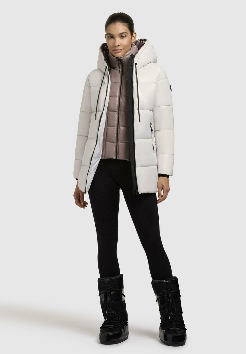 Giubbotto piumino bianco con cappuccio, sovrapposto a una giacca interna marrone trapuntata. Accenti neri e cerniere. Leggings neri e stivali neri lucidi.