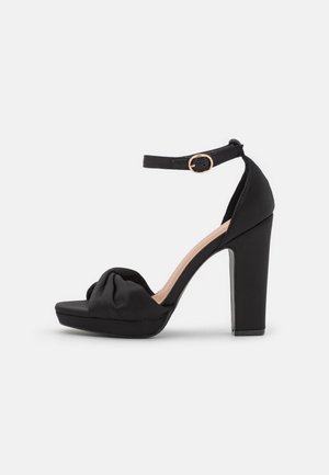 Schwarze High-Heel-Sandale mit Knöchelriemen, goldener Schnalle, offener Zehenpartie und gerafftem Detail am vorderen Riemen auf schlichtem weißem Hintergrund.