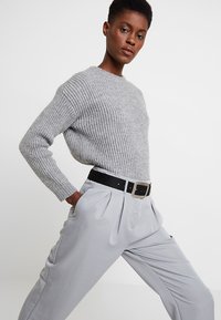 Suéter gris de canalé con mangas largas, combinado con pantalones plisados gris claro. Cinturón negro con hebilla cuadrada de plata.