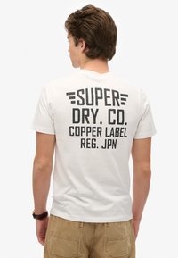 Witte t-shirt met gedurfde zwarte tekst op de achterkant die luidt: "SUPER DRY CO. COPPER LABEL REG. JPN." Korte mouwen en een ronde hals ontwerp.