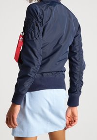 Granatowa kurtka bombowa o gładkiej, błyszczącej teksturze, z ribbowanymi mankietami i dołem, oraz z czerwoną zawieszką do kluczy przypiętą do rękawa.