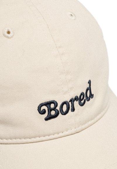 Bežinė medvilninė kepurė su tamsiai mėlynu siuvinėtu užrašu "Bored" priekyje, su išlenktu kraštu ir matomais siuvimo detalėmis.