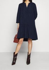 Marinblå bomullsklänning med V-ringning, trekvartsärmar och en lager-på-lager kjol. I kombination med vinröda knähöga stövlar och en texturerad röd handväska.