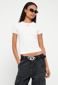 Balta īsroku rāvējaciska T-krekls ar nelielu emblēmu krūšu rajonā, kopā ar melnām kravas biksēm un melnu jostu ar sudraba sprādzi.