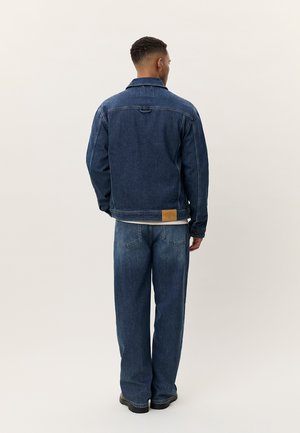 Άνδρας που φοράει σκούρο μπλε τζιν μπουφάν και τζιν παντελόνι στο ίδιο χρώμα, στέκεται με γυρισμένη την πλάτη στην κάμερα, μπροστά σε φωτεινό φόντο.