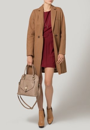 Donna con indosso un cappotto cammello sopra un vestito bordeaux, che tiene una borsa beige e indossa stivaletti color cuoio su uno sfondo neutro.