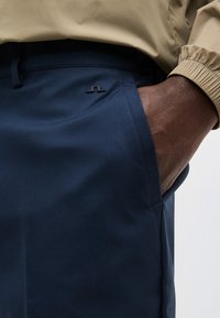 Σκούρο δέρμα χεριού στην τσέπη μπλε ναυτικού παντελονιού με μικρό λογότυπο, μανίκι μπεζ μπουφάν ορατό πάνω από τον καρπό, απλό ανοιχτό φόντο.