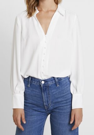 Vrouw draagt een wit blouse met knopen en lange mouwen en blauwe jeans met hoge taille, staand met ontspannen armen.