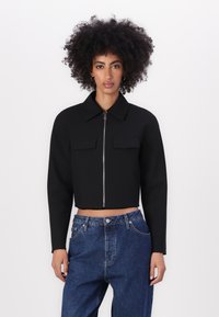Zwarte cropped jacket met een kraag, sluiting met ritssluiting aan de voorkant en twee borstzakken. G gedragen met high-waisted blauwe spijkerbroek.