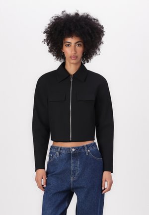Zwarte cropped jacket met een kraag, sluiting met ritssluiting aan de voorkant en twee borstzakken. G gedragen met high-waisted blauwe spijkerbroek.