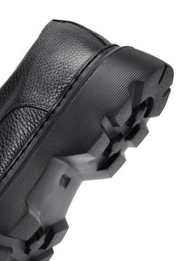 Chaussure en cuir noir avec un dessus texturé, semelle en caoutchouc présentant des crampons prononcés, et un dos légèrement surélevé pour plus de stabilité.
