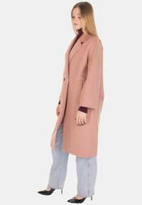 Cappotto lungo color pesca con silhouette aderente, chiusura con un bottone, ampi revers e tasche laterali, abbinato a jeans larghi blu a gamba larga.