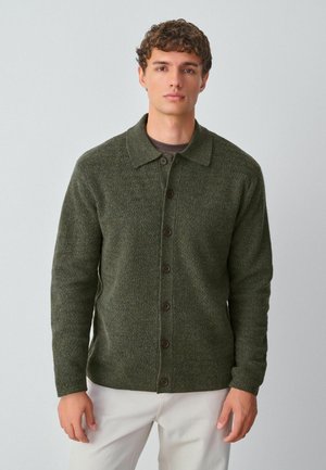 Grüne Wolljacke mit Kragen und Knopfverschluss, strukturierter Stoff und langen Ärmeln, kombiniert mit cremefarbenen Hosen.