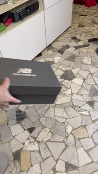 Main tenant une boîte à chaussures New Balance grise au-dessus d'un sol en carrelage mosaïque à côté de placards blancs et d'une étagère avec une console Xbox.