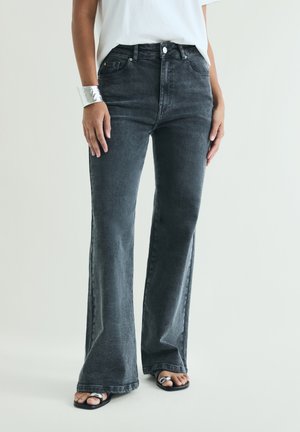 Person wearing dark gray flared jeans, white top, silver cuff bracelet, and black open-toe sandals with a metallic detail.