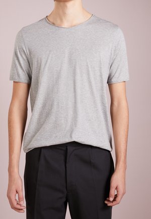 Personne portant un t-shirt à manches courtes gris clair uni, rentré dans un pantalon noir, debout devant un fond mauve clair.