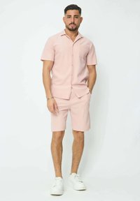 Chemise à boutons à manches courtes rose clair avec une poche poitrine, accompagnée de shorts assortis. Portée avec des baskets blanches, présentant un tissu texturé.